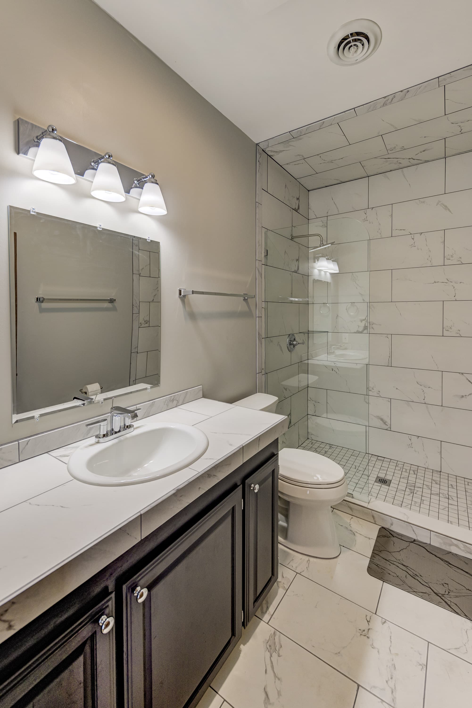 A modern bathroom featuring a glass shower, marble tiles, a dark wood vanity, and bright lighting.