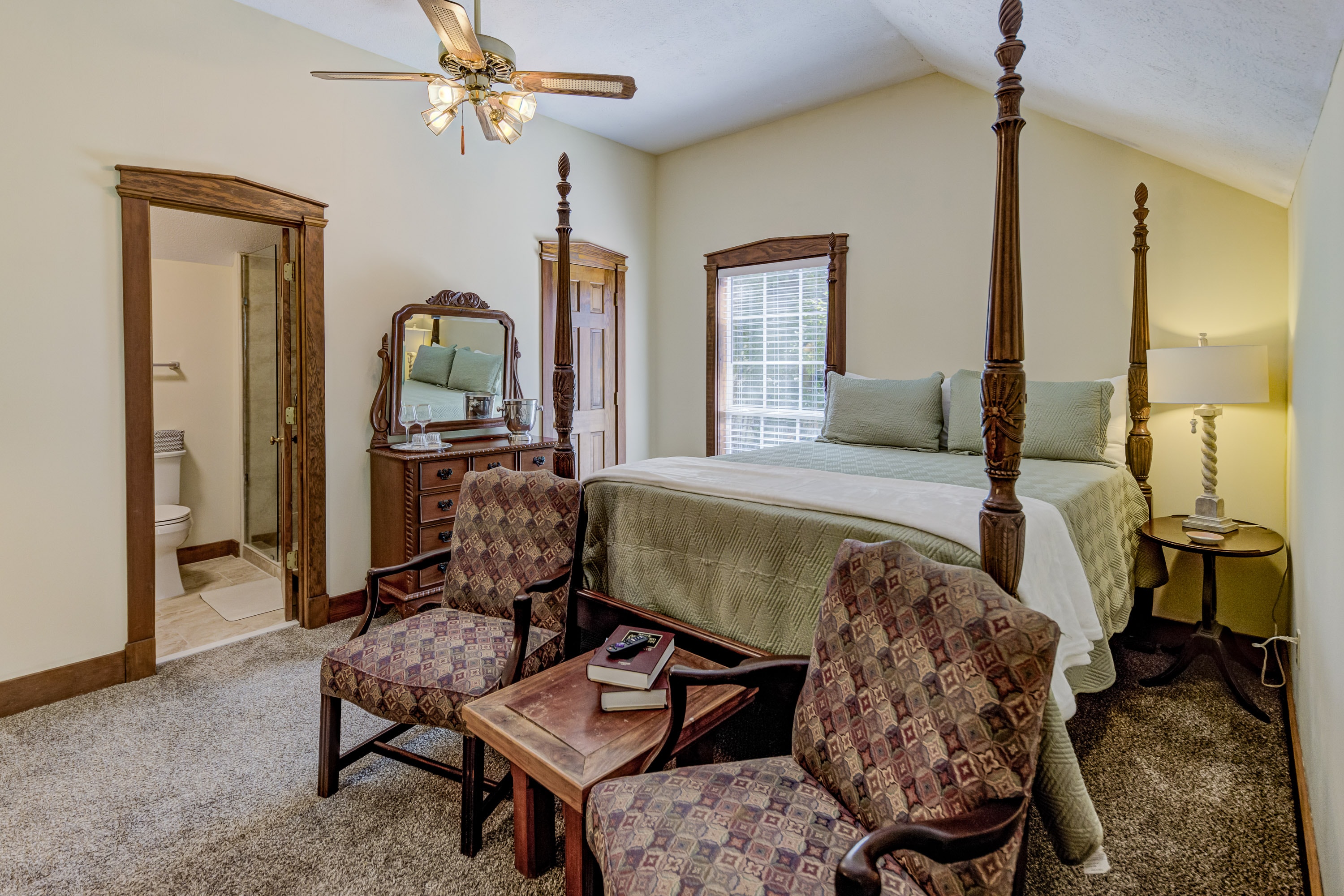A cozy bedroom featuring a queen bed, two armchairs, a wooden dresser, and an en-suite bathroom.
