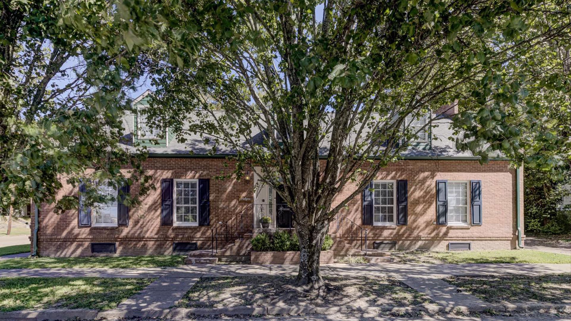 A brick house with multiple windows, framed by a large tree in front.