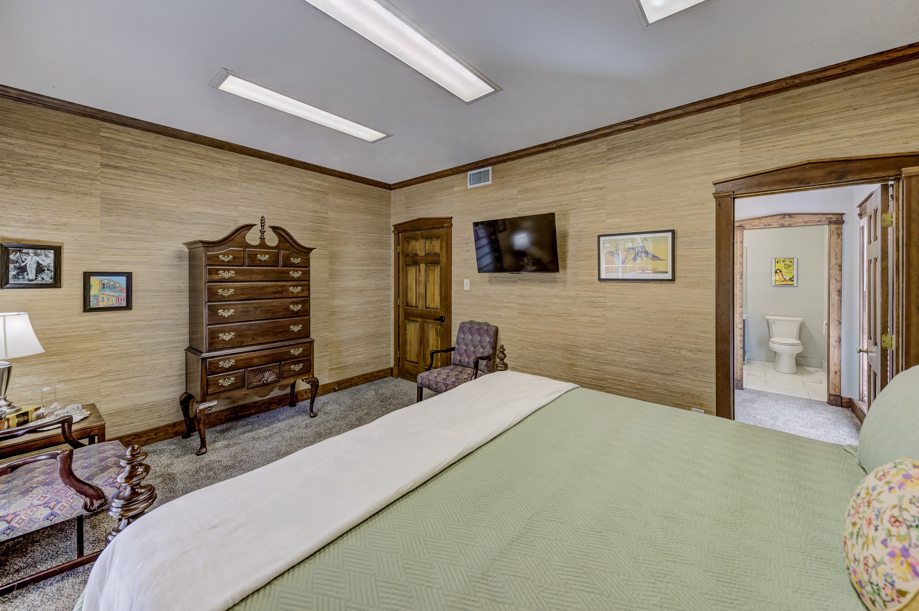 A neatly arranged bedroom featuring a green bedspread, a wooden dresser, a small seating area, and a wall-mounted TV.