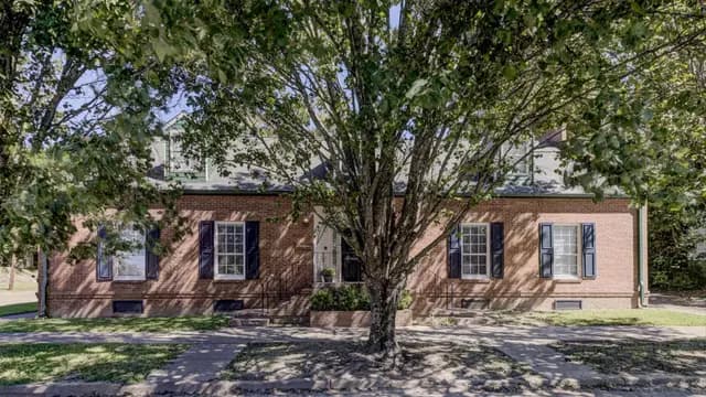 A brick house with dark shutters is partially obscured by a large tree in the front yard.