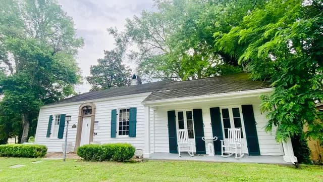 A white house with black shutters and rocking chairs on the porch, surrounded by greenery.