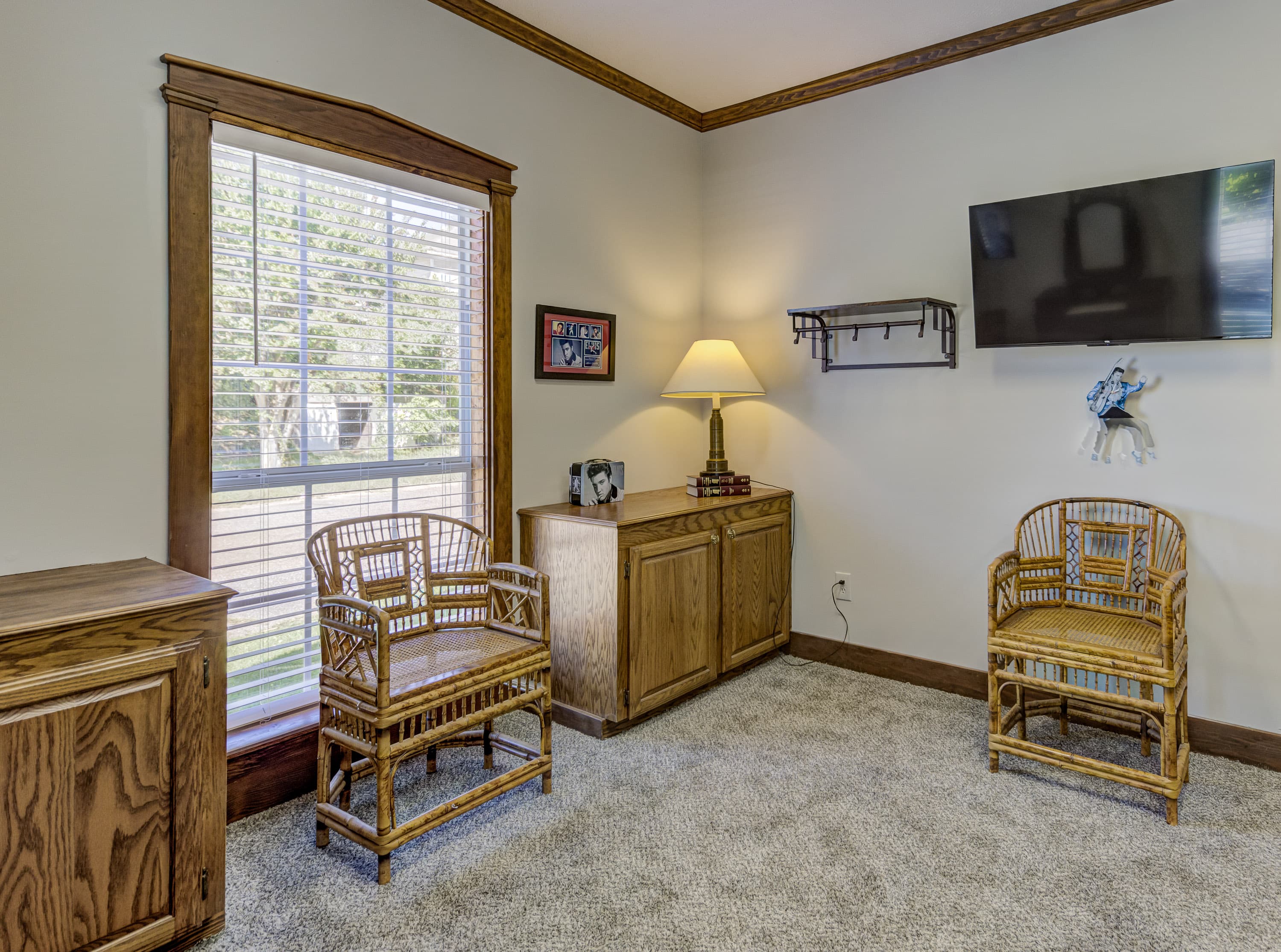 Cozy sitting area featuring two wicker chairs, a lamp, and a wall-mounted TV, with natural light from a window.