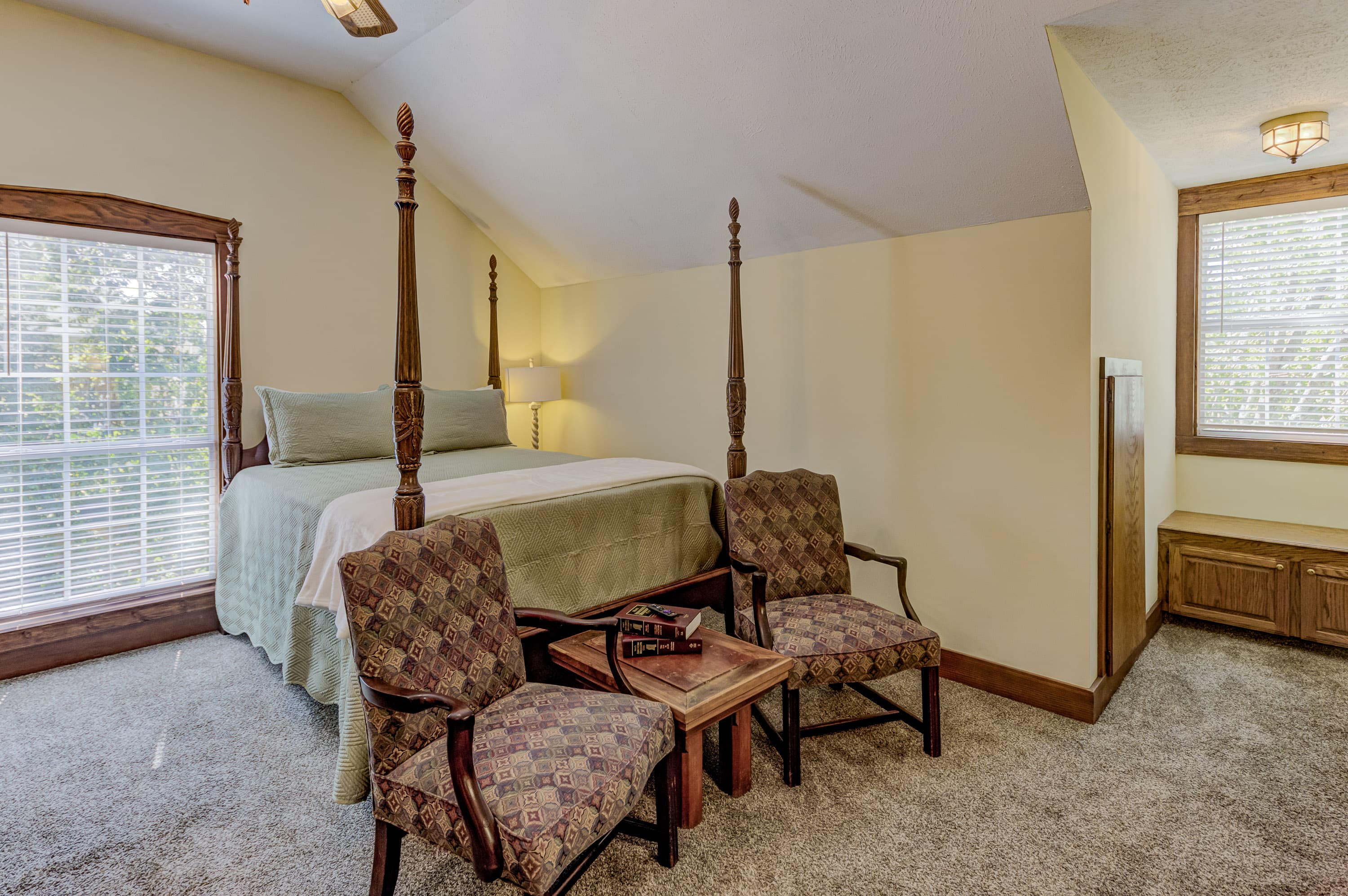 A cozy bedroom featuring a four-poster bed, two upholstered chairs, and natural light from a window.