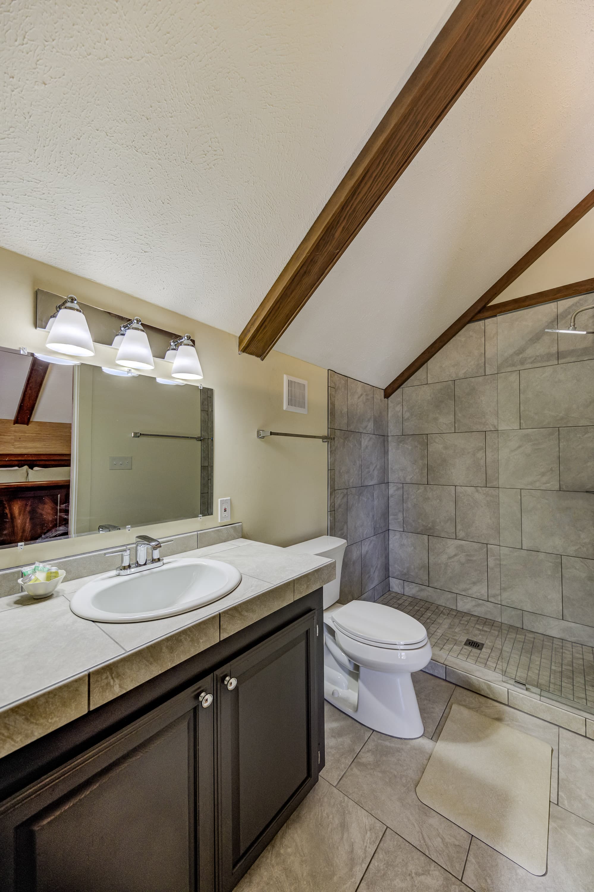 Modern bathroom featuring a sink, toilet, and shower with tiled walls and wooden beams.