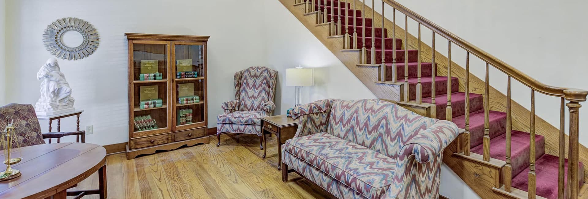 A cozy living area featuring patterned sofas, a glass cabinet, and a staircase.