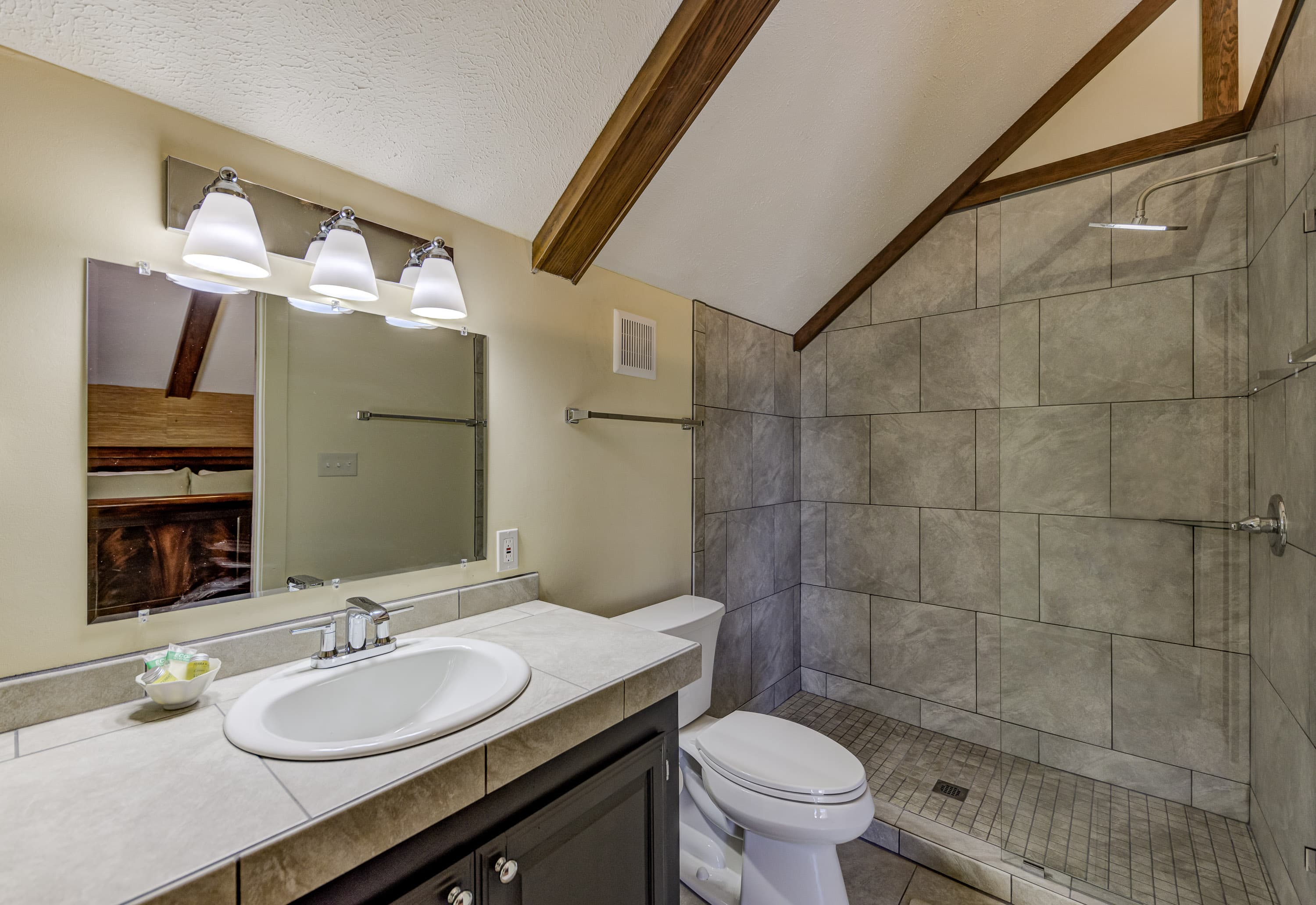 A modern bathroom featuring a sink, toilet, and a walk-in shower with tiled walls and wooden beams.