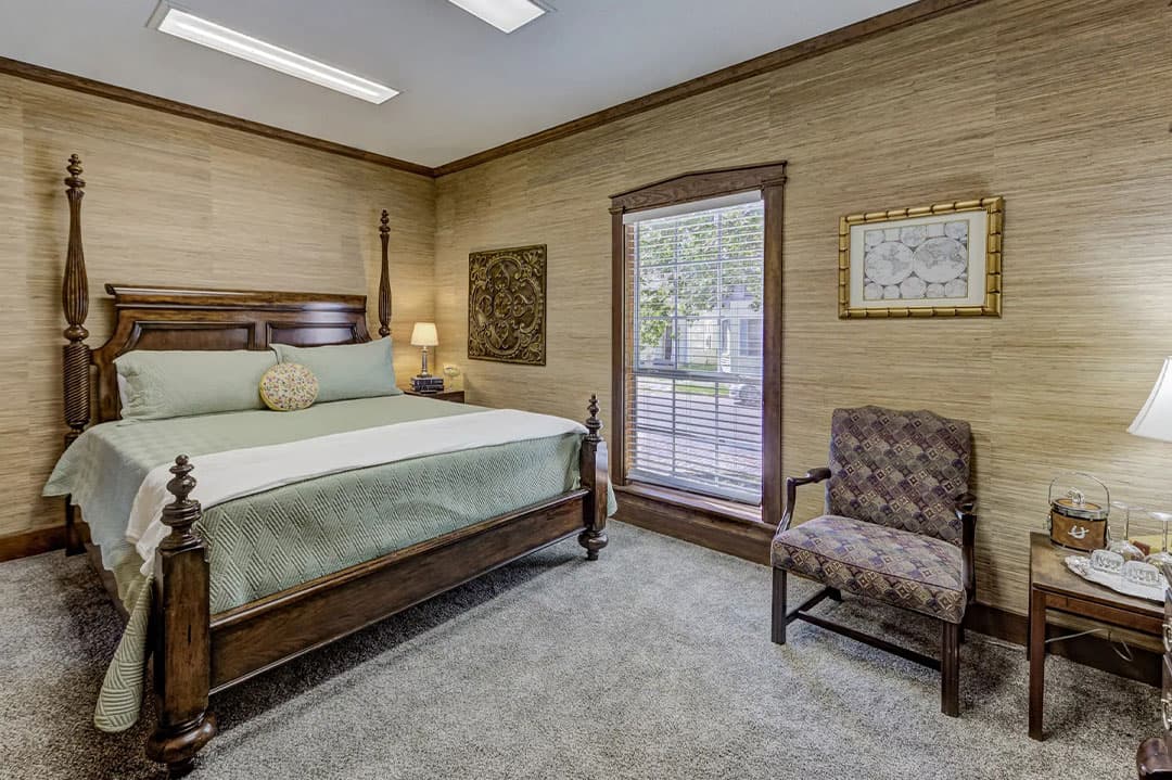 A cozy bedroom featuring a wooden bed, decorative pillows, a patterned armchair, and natural light from a window.